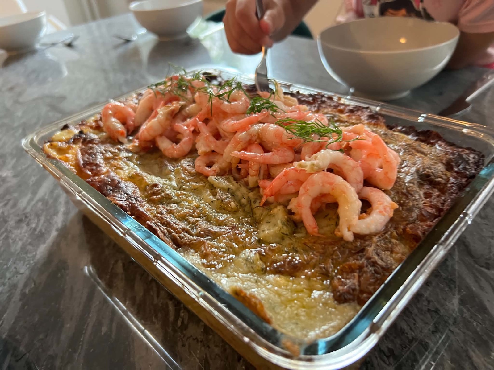 Fiskgratäng på torsk toppad med räkor och dill i en ugnsform, ett barn tar för sig med gaffel